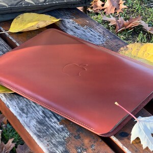May include: A brown leather case with a stitched edge and an Apple logo embossed on the front.