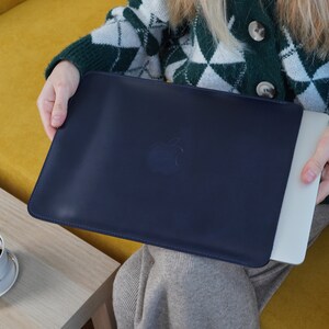 K&ouml;nnte beinhalten: Eine dunkelblaue Laptoptasche mit einem dezenten, gepr&auml;gten Apple-Logo. Die Tasche ist aus einem weichen, strapazierf&auml;higen Material gefertigt und wurde entwickelt, um Ihren Laptop vor Kratzern und St&ouml;&szlig;en zu sch&uuml;tzen.