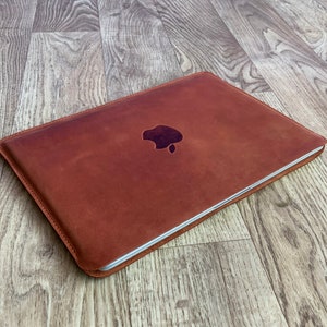 Puede incluir: Una funda para portátil de cuero marrón con un logotipo de Apple en relieve en la parte delantera.