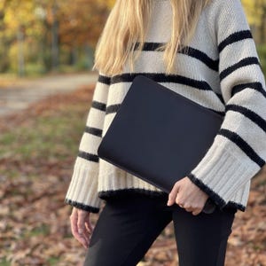 K&ouml;nnte beinhalten: Eine schwarze Laptoptasche wird vor einer Person gehalten, die einen cremefarbenen Pullover mit schwarzen horizontalen Streifen tr&auml;gt. Die &Auml;rmel und der Saum des Pullovers sind schwarz eingefasst. Die Person tr&auml;gt auch eine schwarze Hose.
