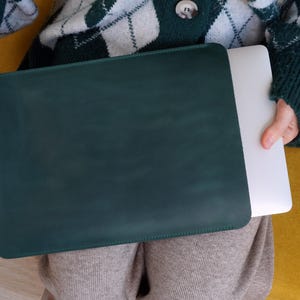 K&ouml;nnte beinhalten: Eine dunkelgr&uuml;ne Laptop-H&uuml;lle mit einem passenden Laptop im Inneren. Die H&uuml;lle ist rechteckig und scheint aus Leder zu sein. Der Laptop ist wei&szlig;. Die Person, die den Laptop h&auml;lt, tr&auml;gt einen gr&uuml;n-wei&szlig;en Argyle-Pullover.