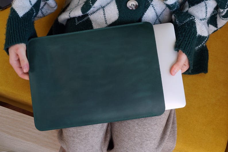 K&ouml;nnte beinhalten: Eine dunkelgr&uuml;ne Laptop-H&uuml;lle mit einem passenden Laptop im Inneren. Die H&uuml;lle ist rechteckig und scheint aus Leder zu sein. Der Laptop ist wei&szlig;. Die Person, die den Laptop h&auml;lt, tr&auml;gt einen gr&uuml;n-wei&szlig;en Argyle-Pullover.