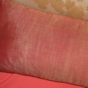 May include: A rectangular, reddish-orange pillow with a textured surface. The pillow is placed on a coral-colored surface, with a floral patterned pillow in the background. The pillow is likely for home decor.