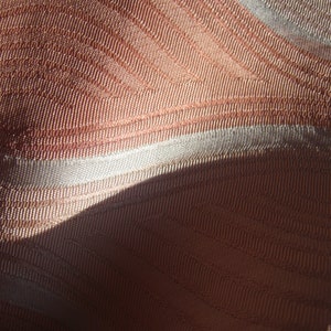 May include: A close-up view of fabric featuring a woven texture with alternating bands of copper-toned and light silver-white threads. The material displays a subtle, repeating wave-like pattern created by the direction of the weave and light.