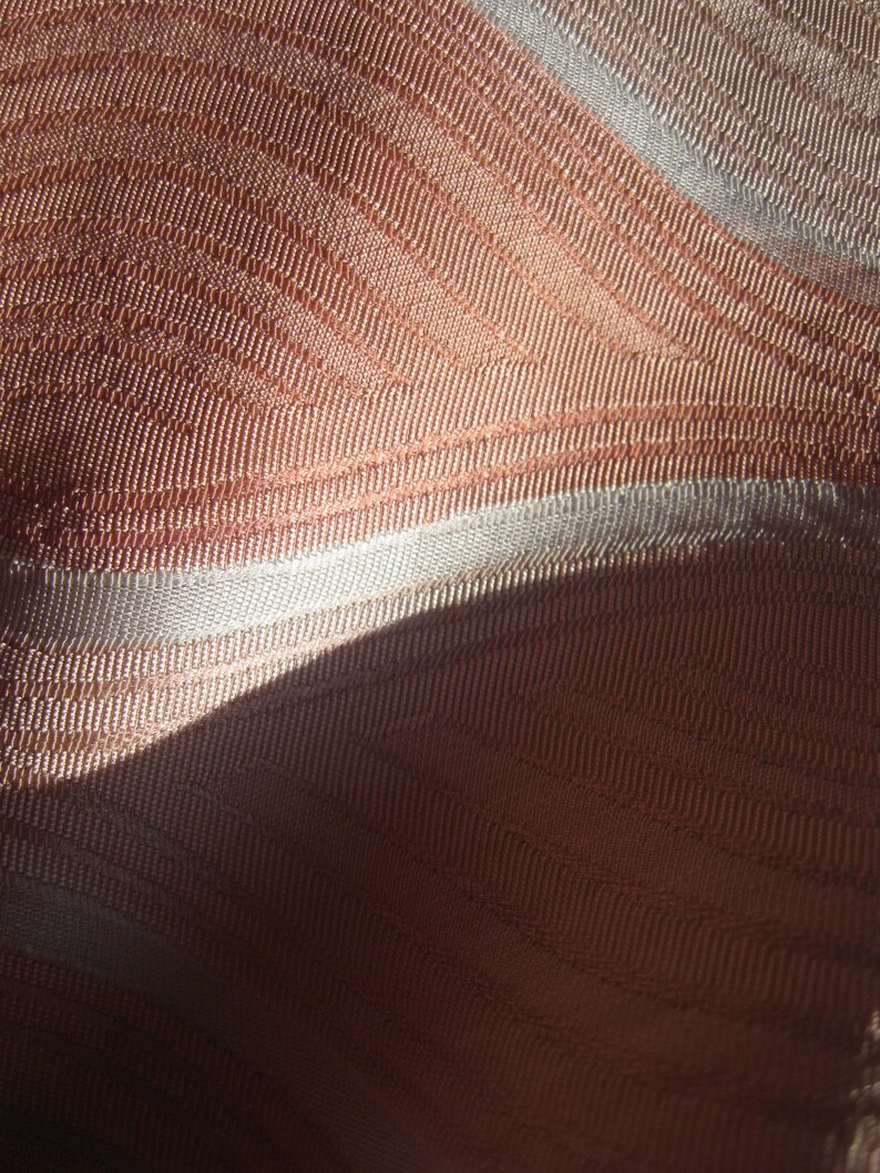 May include: A close-up view of fabric featuring a woven texture with alternating bands of copper-toned and light silver-white threads. The material displays a subtle, repeating wave-like pattern created by the direction of the weave and light.