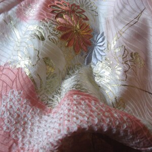 May include: A close-up of a pink and white fabric with gold and silver embroidery. The fabric has a delicate floral pattern with a textured surface.