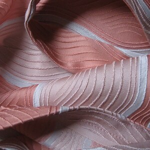 May include: A fabric featuring a repeating pattern of wavy, curved lines in shades of salmon pink and light gray. The material has a textured, ribbed surface with a subtle sheen, creating a series of folds and drapes across the entire frame.