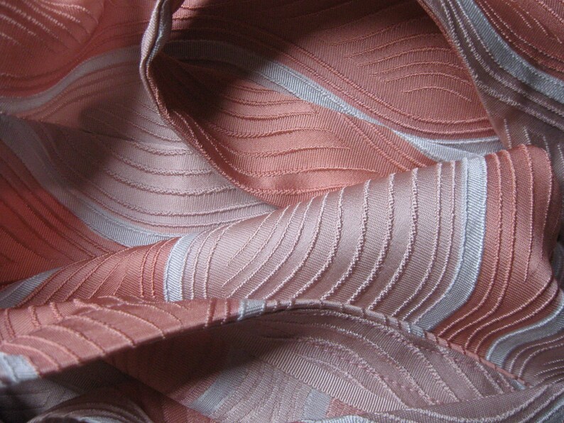 May include: A fabric featuring a repeating pattern of wavy, curved lines in shades of salmon pink and light gray. The material has a textured, ribbed surface with a subtle sheen, creating a series of folds and drapes across the entire frame.