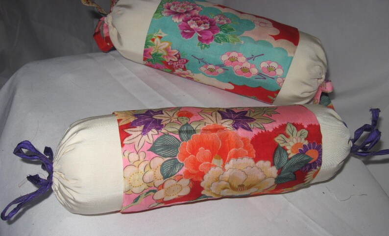 May include: Two white fabric cylindrical pillows with colorful floral patterns. The pillows have purple ties at the ends.