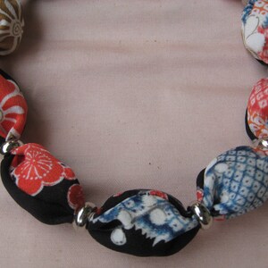 May include: A necklace featuring a series of fabric-covered beads in various patterns and colors, including red, blue, black, and tan, separated by small, smooth, reflective silver-toned metal spacer beads arranged in a circular formation.