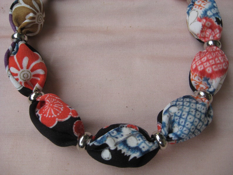 May include: A necklace featuring a series of fabric-covered beads in various patterns and colors, including red, blue, black, and tan, separated by small, smooth, reflective silver-toned metal spacer beads arranged in a circular formation.