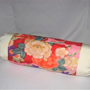 May include: A cylindrical pillow with a white fabric cover and a floral print fabric insert. The floral print features pink, red, orange, yellow, green, and purple flowers on a pink background. The pillow has purple ties at each end.