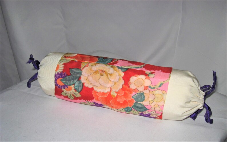 May include: A cylindrical pillow with a white fabric cover and a floral print fabric insert. The floral print features pink, red, orange, yellow, green, and purple flowers on a pink background. The pillow has purple ties at each end.