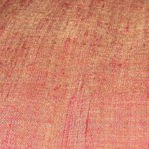 May include: Close-up of a textured fabric with a woven pattern. The fabric has a gradient of colors, transitioning from a reddish-orange hue to a lighter, golden-brown shade. The weave creates a subtle, grid-like texture across the surface.