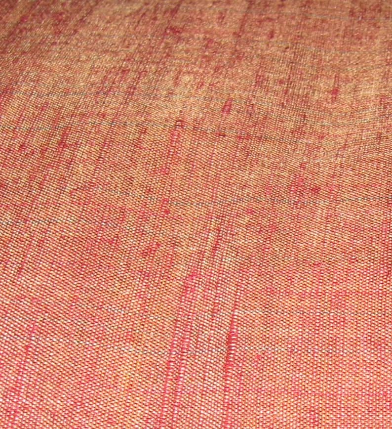 May include: Close-up of a textured fabric with a woven pattern. The fabric has a gradient of colors, transitioning from a reddish-orange hue to a lighter, golden-brown shade. The weave creates a subtle, grid-like texture across the surface.