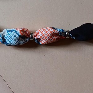 May include: A bracelet featuring a series of fabric-covered beads in various patterns and colors, including red, blue, black, and orange, separated by small silver-toned metal spacers, finished with a circular metal clasp at one end.
