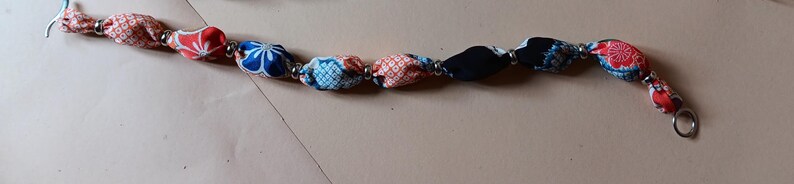 May include: A bracelet featuring a series of fabric-covered beads in various patterns and colors, including red, blue, black, and orange, separated by small silver-toned metal spacers, finished with a circular metal clasp at one end.
