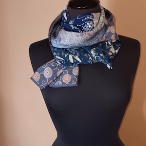 Puede incluir: Bufanda azul marino con estampados florales en tonos blancos y melocotón, drapeada sobre un maniquí negro. La bufanda tiene una variedad de patrones y texturas, incluyendo una sección con un diseño azul oscuro y blanco. Accesorio de moda.