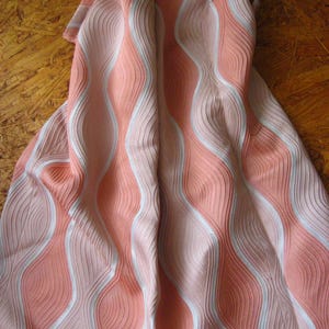 May include: A fabric textile featuring a repeating vertical wave pattern in shades of coral pink and white. The material has a distinct ribbed or pleated texture that creates a sense of depth and movement across the surface of the folded cloth.