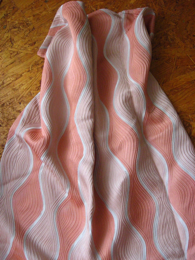 May include: A fabric textile featuring a repeating vertical wave pattern in shades of coral pink and white. The material has a distinct ribbed or pleated texture that creates a sense of depth and movement across the surface of the folded cloth.
