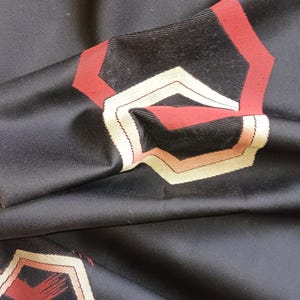 Könnte beinhalten: Ein schwarzer Stoff mit einem sich wiederholenden geometrischen Muster aus ineinander verschachtelten Sechsecken in den Farben Dunkelrot, Creme und Hellbraun. Das Material hat eine glatte, leicht glänzende Oberfläche mit sichtbaren Falten.
