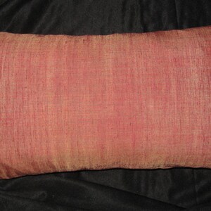 May include: A rectangular decorative pillow with a reddish-pink hue. The pillow has a textured surface and appears to be made of a silky fabric. The pillow is placed on a dark background.