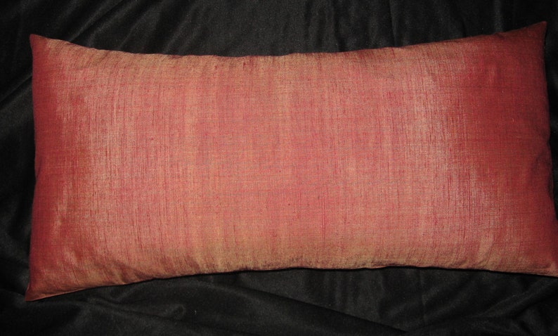 May include: A rectangular decorative pillow with a reddish-pink hue. The pillow has a textured surface and appears to be made of a silky fabric. The pillow is placed on a dark background.