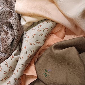 Könnte beinhalten: Eine Ansammlung gefalteter Stoffstücke in verschiedenen Texturen und Farben, darunter ein blasspfirsichfarbenes Textil mit Blumenmuster, ein dunkelbrauner Stoff mit gesprenkeltem Design, ein hellcremefarbener Stoff mit Blattmotiven und ein texturiertes olivgrünes Stück.