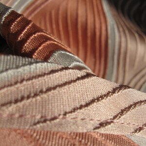 May include: A close-up view of fabric featuring a series of raised, parallel ridges in alternating shades of copper, bronze, and light beige. The material displays a distinct woven texture with a subtle sheen, creating a rhythmic pattern of light and shadow across the surface of the textile.