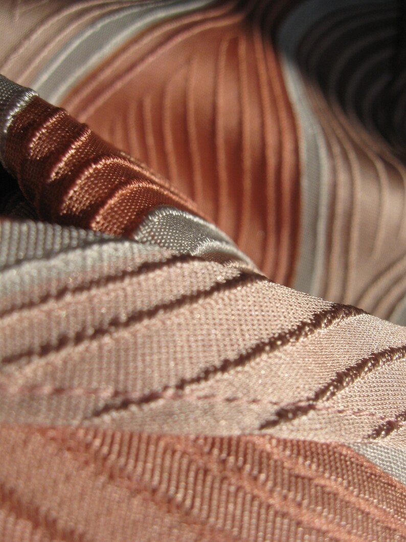 May include: A close-up view of fabric featuring a series of raised, parallel ridges in alternating shades of copper, bronze, and light beige. The material displays a distinct woven texture with a subtle sheen, creating a rhythmic pattern of light and shadow across the surface of the textile.