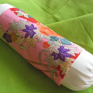 May include: A white and pink cylindrical pillow with a floral pattern. The pillow has a white fabric on the ends and a pink fabric with a floral pattern in the middle. The pillow is lying on a green surface.