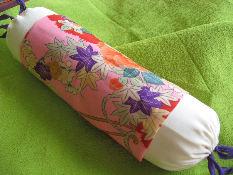 May include: A white and pink cylindrical pillow with a floral pattern. The pillow has a white fabric on the ends and a pink fabric with a floral pattern in the middle. The pillow is lying on a green surface.