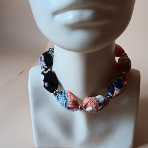 May include: A necklace featuring fabric-covered beads in various patterns and colors, including black, orange, blue, and white, separated by small silver-toned metallic spacer beads. The necklace is displayed on a white mannequin bust.