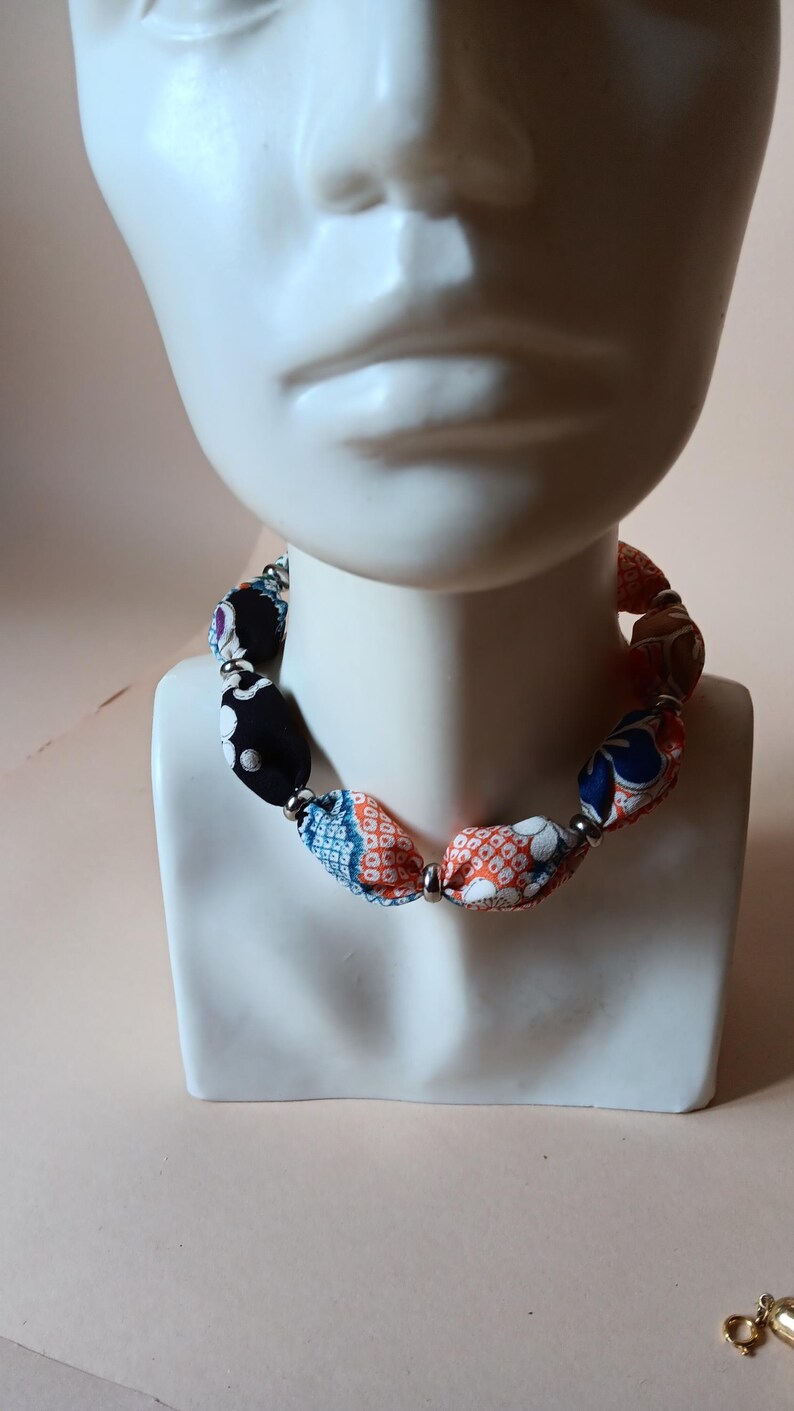 May include: A necklace featuring fabric-covered beads in various patterns and colors, including black, orange, blue, and white, separated by small silver-toned metallic spacer beads. The necklace is displayed on a white mannequin bust.