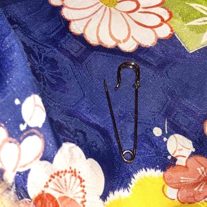 Könnte beinhalten: Eine dunkelmetallische Sicherheitsnadel auf einem blauen Stoffhintergrund mit einem sich wiederholenden Muster aus weißen, rosa und gelben Blüten. Die Sicherheitsnadel ist geöffnet und zeigt ihre scharfe Nadelspitze und den Federungsmechanismus.