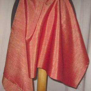 May include: A draped piece of red-orange fabric with a subtle sheen. The fabric is draped over a black form and a wooden stand, showcasing its texture and color. The fabric's edges are visible, highlighting its construction.