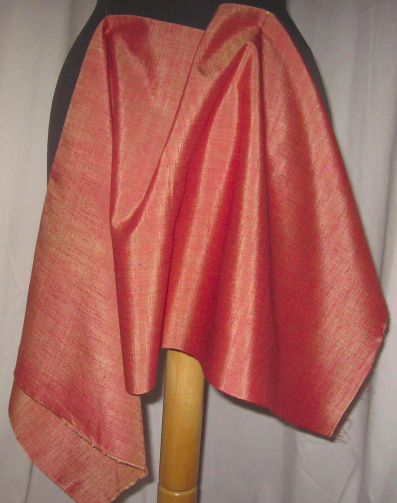 May include: A draped piece of red-orange fabric with a subtle sheen. The fabric is draped over a black form and a wooden stand, showcasing its texture and color. The fabric's edges are visible, highlighting its construction.