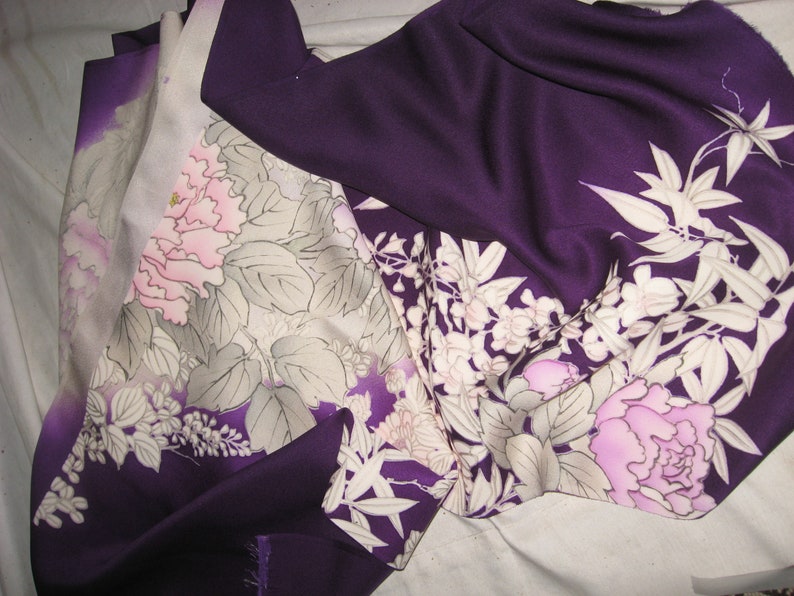 May include: Close-up of a folded fabric with a floral pattern. The fabric is primarily deep purple with a gradient to lighter shades. The floral design features pink and white flowers with gray leaves.