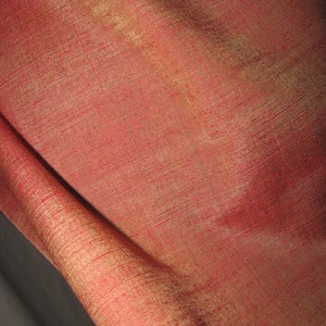 May include: Close-up of a luxurious red fabric with a subtle sheen. The material has a textured appearance with visible threads, and a golden hue is visible in the folds. The fabric is likely silk or a similar high-quality textile.