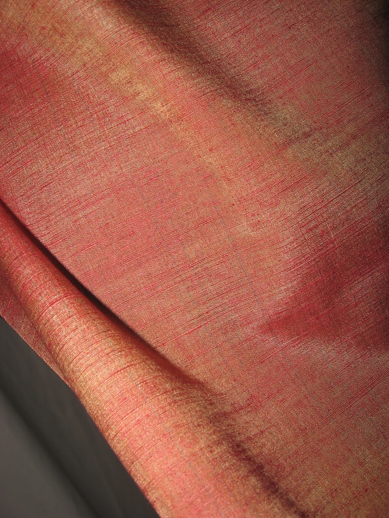 May include: Close-up of a luxurious red fabric with a subtle sheen. The material has a textured appearance with visible threads, and a golden hue is visible in the folds. The fabric is likely silk or a similar high-quality textile.