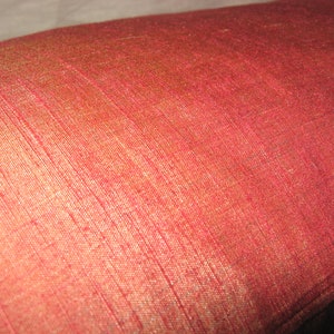 May include: Close-up of a red pillow with a textured, woven fabric. The pillow has a slight sheen, with variations in color from red to a golden hue. The fabric appears to be silk or a similar material.