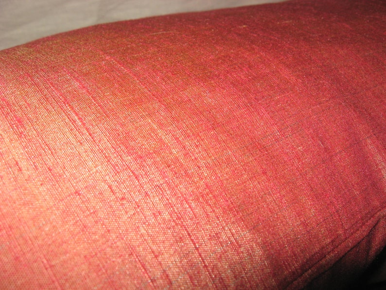May include: Close-up of a red pillow with a textured, woven fabric. The pillow has a slight sheen, with variations in color from red to a golden hue. The fabric appears to be silk or a similar material.
