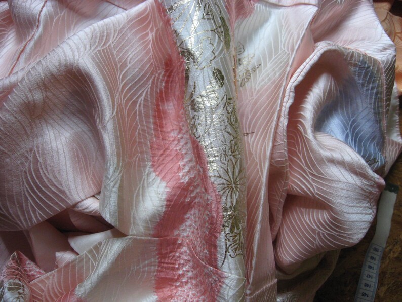 May include: A pink and white silk fabric with a gold floral pattern. The fabric is folded and draped, showing the texture and sheen of the material.