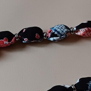 May include: A necklace featuring a series of fabric-covered beads in black, red, white, and blue patterns, separated by small metallic silver spacers. The beads are strung together in a continuous line, showcasing various floral and geometric textile designs.