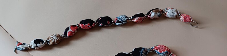 May include: A necklace featuring a series of fabric-covered beads in black, red, white, and blue patterns, separated by small metallic silver spacers. The beads are strung together in a continuous line, showcasing various floral and geometric textile designs.