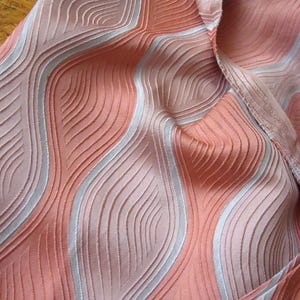 May include: A piece of fabric featuring a textured, wavy pattern in shades of peach, coral, and light pink. The design consists of raised, vertical, undulating lines that create a sense of depth and movement across the surface of the material.