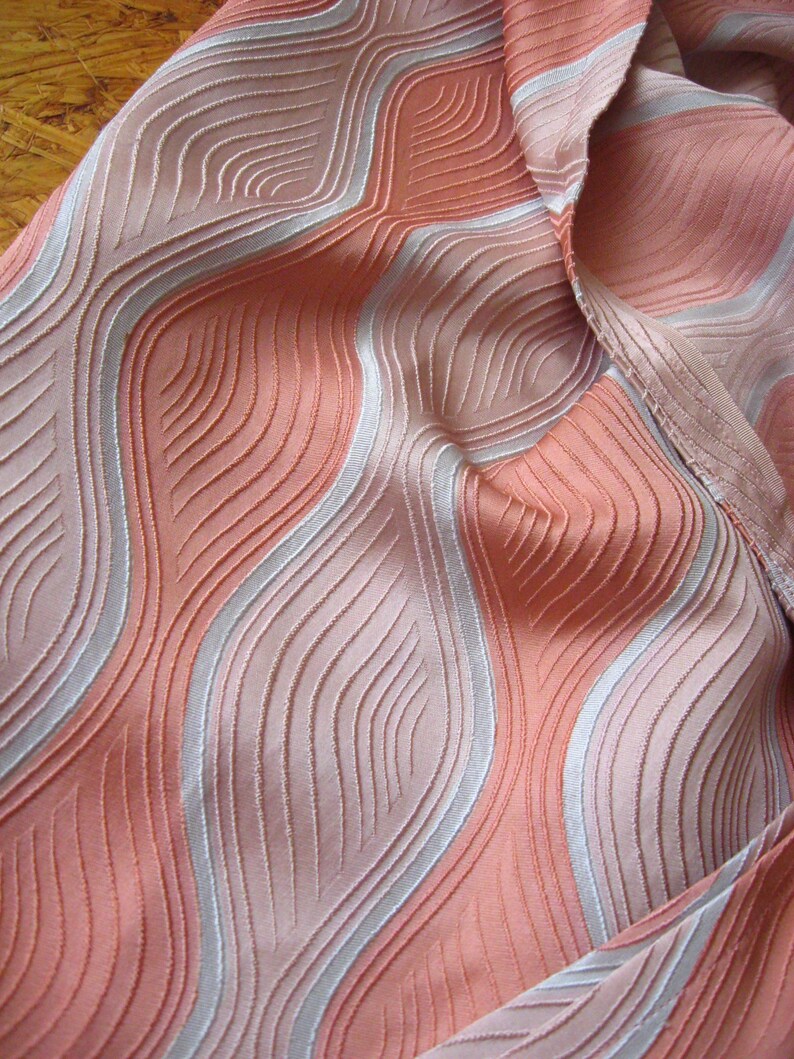 May include: A piece of fabric featuring a textured, wavy pattern in shades of peach, coral, and light pink. The design consists of raised, vertical, undulating lines that create a sense of depth and movement across the surface of the material.
