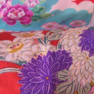 May include: Red fabric with a floral pattern featuring white, purple, and yellow flowers. The fabric has a textured surface.