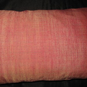 May include: A rectangular decorative pillow with a textured, reddish-pink fabric. The pillow's surface has a subtle sheen, and the color varies slightly across the surface. The pillow is a solid color with no visible patterns.