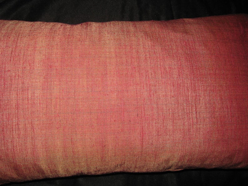 May include: A rectangular decorative pillow with a textured, reddish-pink fabric. The pillow's surface has a subtle sheen, and the color varies slightly across the surface. The pillow is a solid color with no visible patterns.